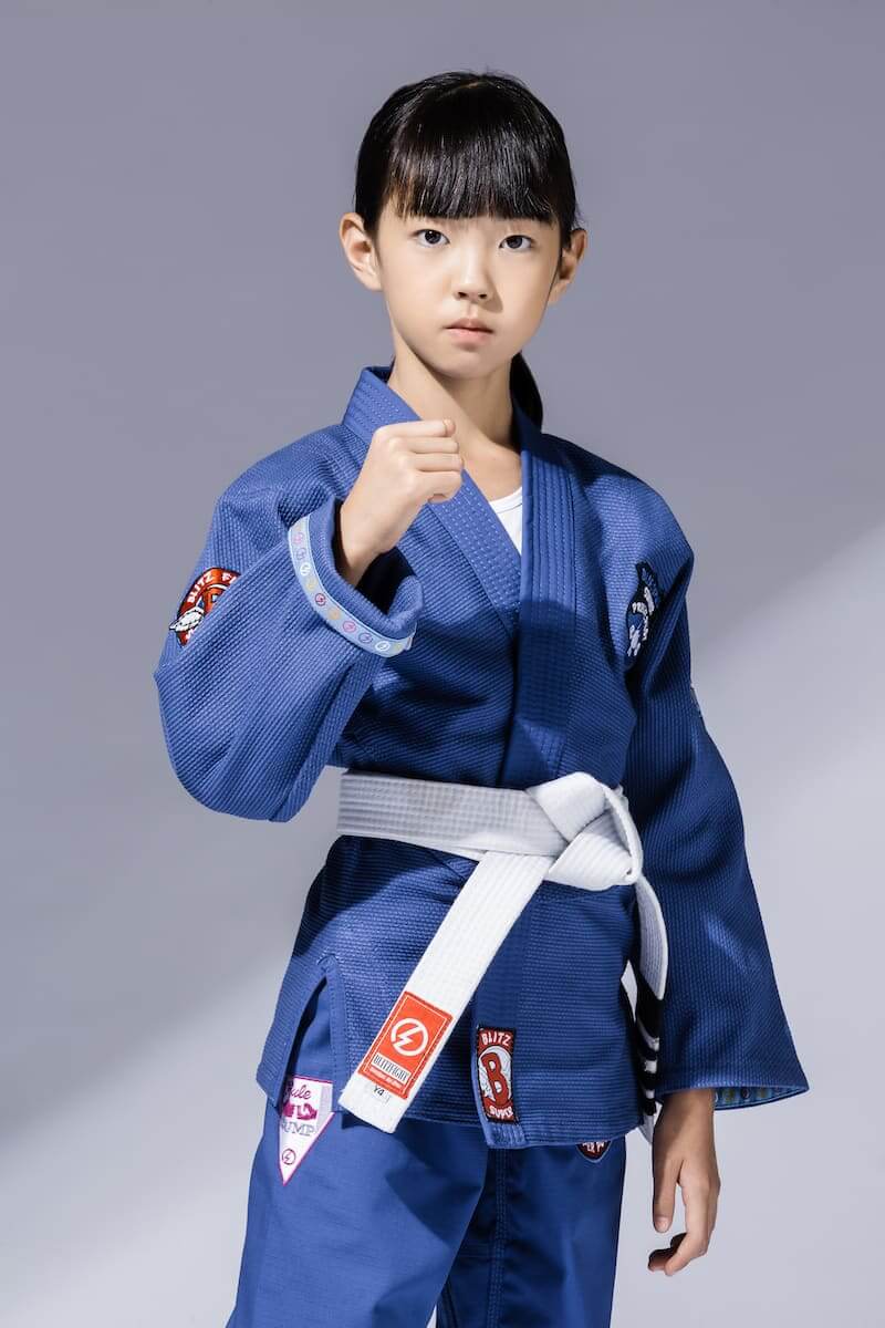 A young girl wearing the BLITZ Kids Super Power BJJ Gi poses confidently with a white belt.